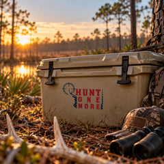 Hunt One More American flag hunting decal on a tan cooler with antlers, binoculars and camo hat at sunrise