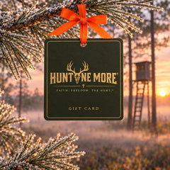 Hunt One More gift card hanging from a frosted pine branch with an orange ribbon at deer camp