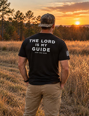 Man wearing the black The Lord Is My Guide® tee, white back print, standing in a golden field at sunset – Hunt One More