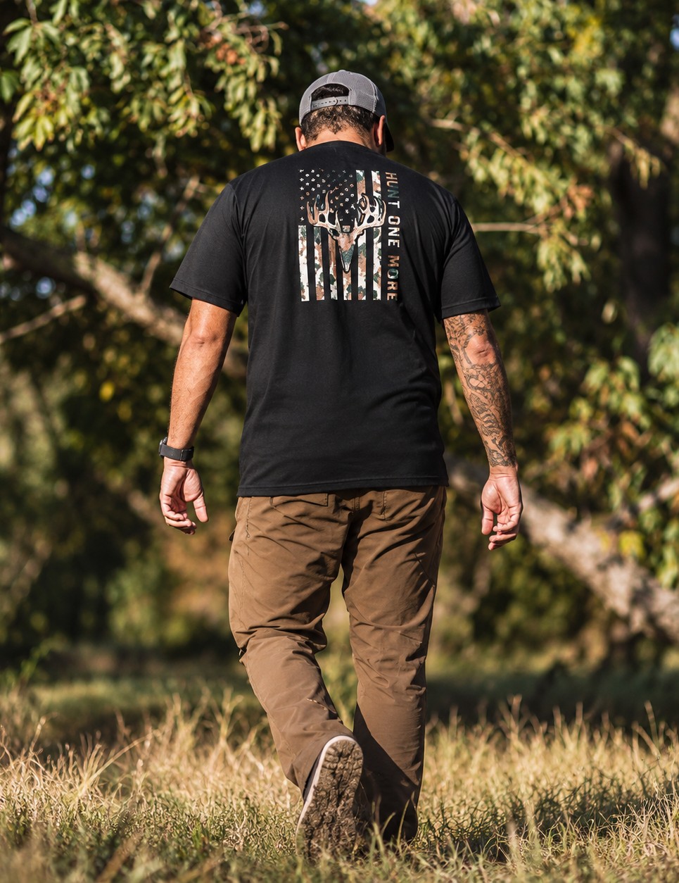 Man wearing the black Stripes and Antlers Camo tee, camo American flag and buck skull back print, walking through the woods – Hunt One More