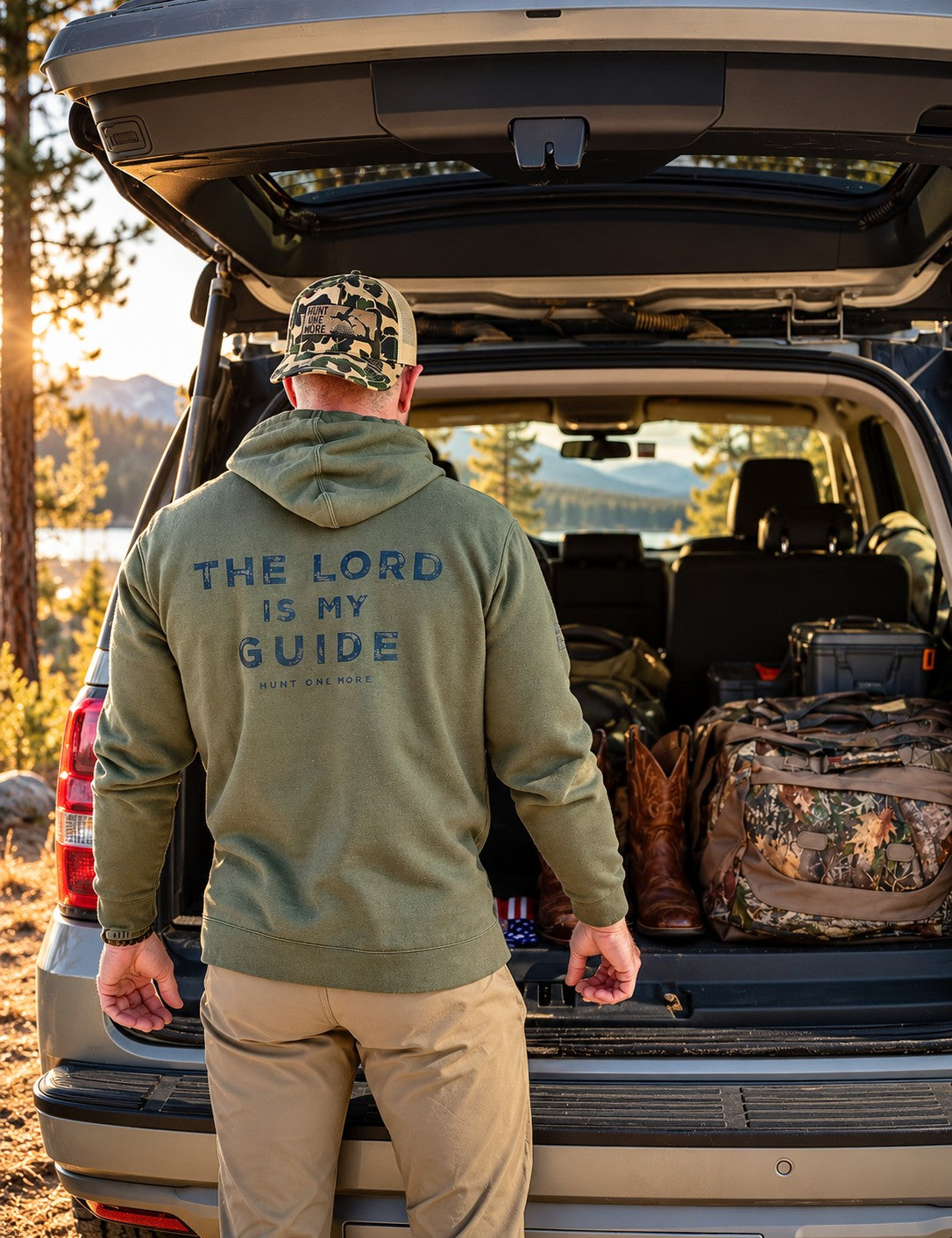 Man wearing the Military Green The Lord Is My Guide® hoodie, back print, loading hunting gear at the truck tailgate – Hunt One More