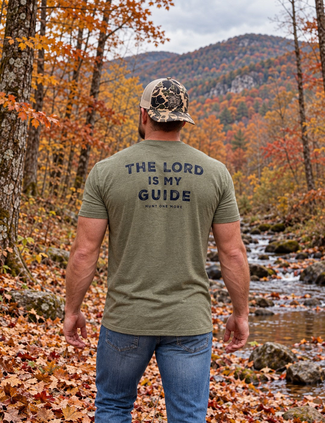 Man wearing the Military Green The Lord Is My Guide® tee, navy back print, beside a creek in autumn hardwoods – Hunt One More