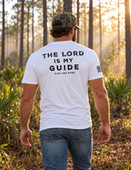 Man wearing the white The Lord Is My Guide® tee, navy back print, walking through a misty pine forest at sunrise – Hunt One More